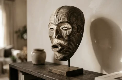 A carved African mask with textured patterns and open mouth is displayed on a wooden stand, placed on a dark shelf next to a ceramic pot, in a softly lit room.