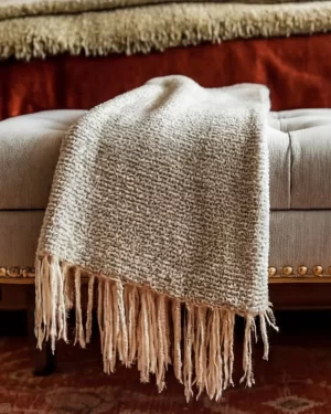 A close-up shot of a textured, cream-colored knit throw blanket with long fringe draped over a grey tufted ottoman. In the blurred background, the rich orange and olive tones of the bedding are visible.