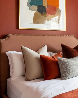 A detail shot of a bed's headboard against a burnt orange wall. Above the studded tan headboard hangs a modern abstract painting in a gold frame featuring blocks of teal, orange, and tan.