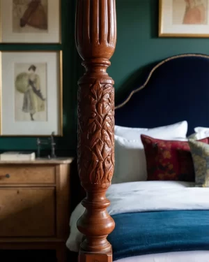 A macro shot focusing on the intricate carvings of a dark wood four-poster bed column. In the soft-focus background, framed vintage sketches of dandy figures hang on a dark green wall.