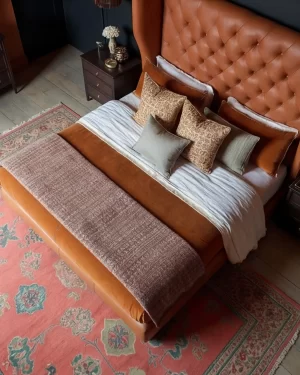 An overhead view of a bed with a large, tufted tan leather headboard and rust-colored bedding. The room features a vibrant pink and green patterned rug over light wood floors.