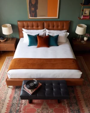 A symmetrical view of a brown leather bed with a black tufted bench at the foot. A vintage record player sits on the bench atop a red Persian rug. Large abstract art in orange and tan hangs centered above the headboard.