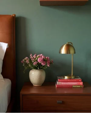 A close-up detail of a mid-century wood nightstand featuring a gold dome-style lamp, a stack of red and pink books, and a speckled cream vase filled with pink carnations against a teal wall.