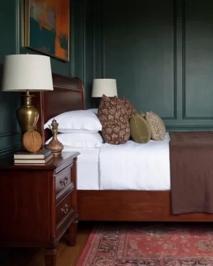 A side view of a dark wood nightstand featuring a gold trophy-style lamp, vintage books, and an antique brass kettle. The bed is layered with white linens and brown floral-patterned cushions.