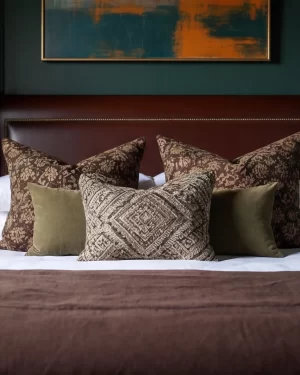 A close-up of textured bedding, featuring large brown floral-print cushions and a center geometric-patterned pillow in muted earth tones against a dark leather headboard.