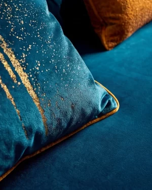 A close-up, angled shot of a plush teal velvet pillow featuring metallic gold splatter patterns, resting on a matching teal sofa.